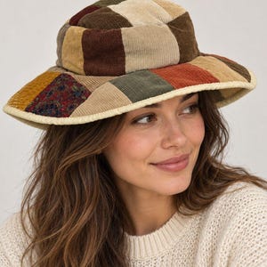 May include: A patchwork bucket hat constructed from corduroy fabric panels in shades of brown, tan, olive green, and rust orange. The hat features a wide, downward-sloping brim with a cream-colored piped edge and a multi-colored floral print patch.