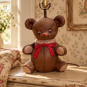 Puede incluir: Una base de lámpara de mesa de cerámica con forma de oso de peluche marrón, con un lazo rojo de lunares, un cuello blanco con detalles de costura rojos y un casquillo de latón que sobresale de la parte superior de la cabeza. El oso tiene pelaje texturizado.