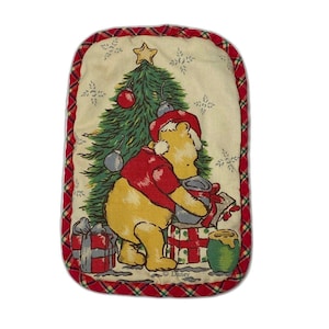 May include: Rectangular Christmas-themed pot holder featuring Winnie the Pooh in a Santa hat, with a Christmas tree and presents. The border is red plaid. The background is cream-colored with snowflake accents. The Disney logo is visible.