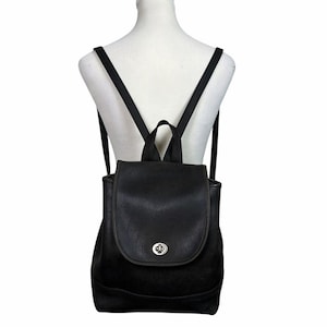 May include: A black leather backpack featuring a fold-over flap with a silver-toned metal turn-lock closure. The bag includes a top handle and two adjustable shoulder straps, showcasing a structured design with a smooth finish and clean lines.