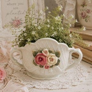 May include: A white ceramic planter with two handles and a lattice-textured surface, featuring a central decorative cluster of sculpted pink and cream roses with green leaves, filled with small green sprigs and tiny white flowers.
