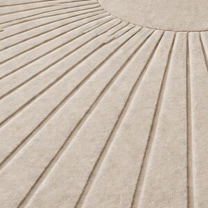 May include: Close-up of a beige rug with a sunburst design. The rug features a circular center and radiating lines, creating a textured pattern. The neutral color and geometric design make it suitable for various interior styles.