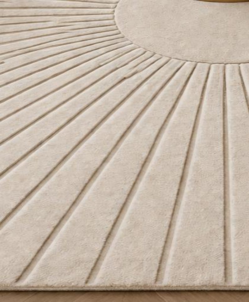 May include: Close-up of a beige rug with a sunburst design. The rug features a circular center and radiating lines, creating a textured pattern. The neutral color and geometric design make it suitable for various interior styles.