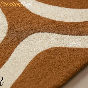 May include: Close-up of a rug with a wavy pattern in shades of brown and cream. The rug has a textured surface and the text "Modern Creation Rugs" is visible in the upper left corner. The letters "MCR" are in the lower left and center right.
