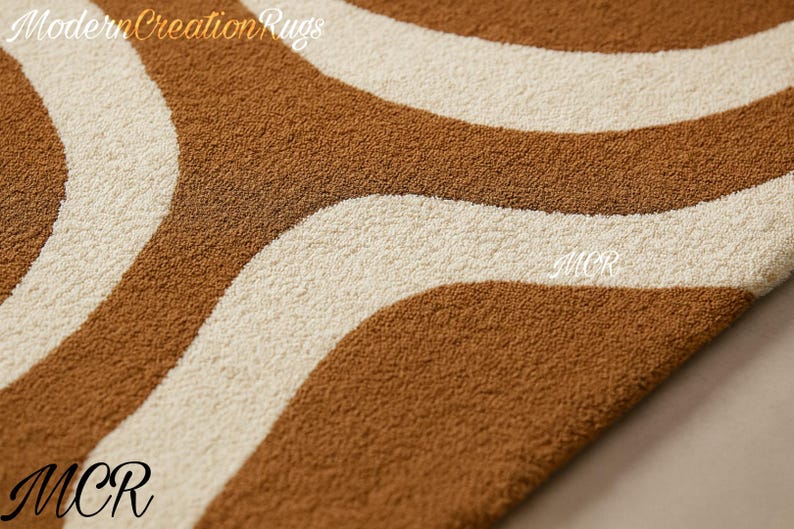 May include: Close-up of a rug with a wavy pattern in shades of brown and cream. The rug has a textured surface and the text "Modern Creation Rugs" is visible in the upper left corner. The letters "MCR" are in the lower left and center right.