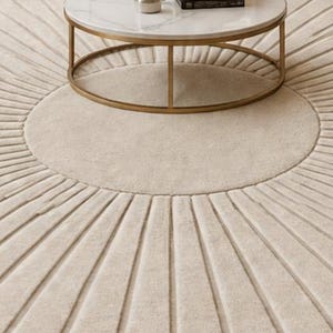 May include: A round, cream-colored rug with a sunburst design. The rug features a central circle surrounded by radiating lines, creating a textured pattern. A gold-framed, marble-topped coffee table sits atop the rug.