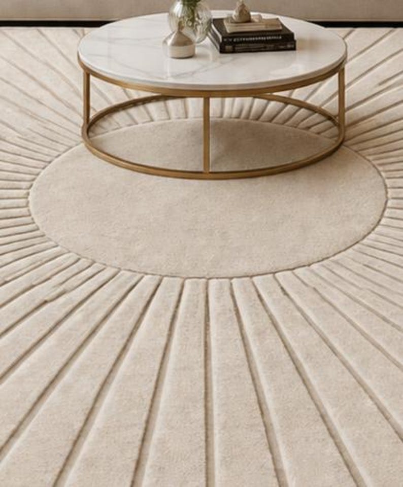 May include: A round, cream-colored rug with a sunburst design. The rug features a central circle surrounded by radiating lines, creating a textured pattern. A gold-framed, marble-topped coffee table sits atop the rug.
