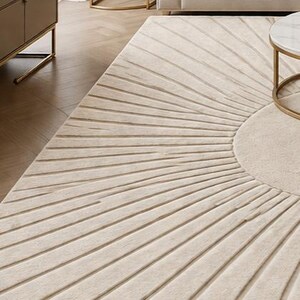 May include: Beige area rug with a sunburst design. The rug features a circular center and radiating lines, creating a textured pattern. The rug is placed in a living room setting, with a sofa and side table visible.