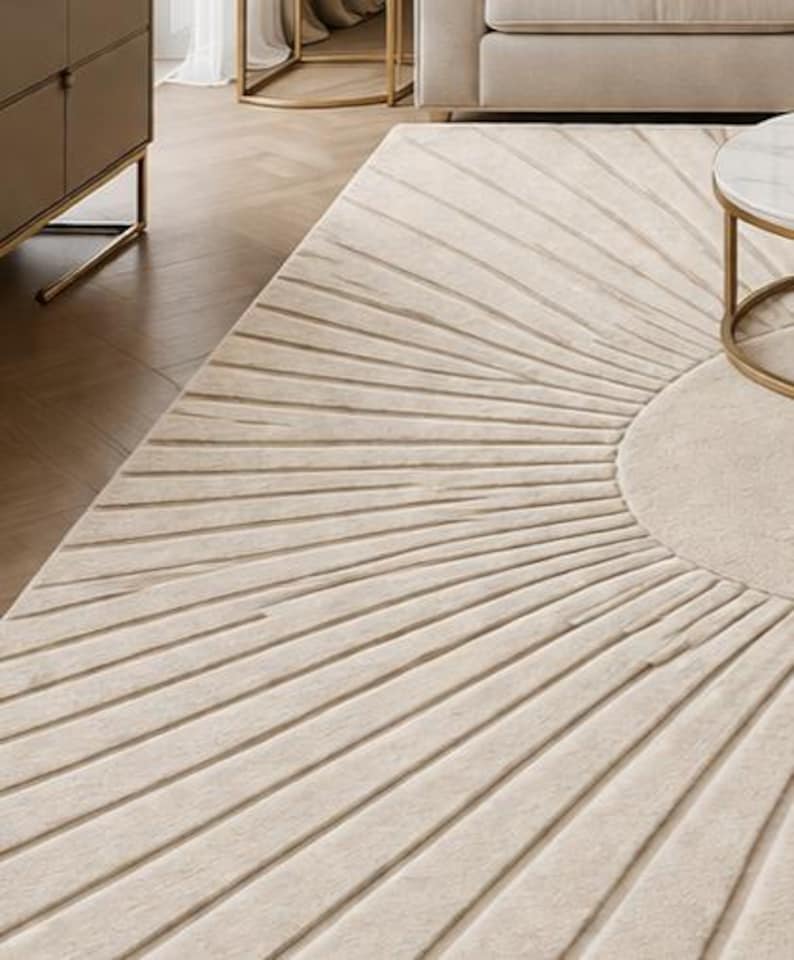 May include: Beige area rug with a sunburst design. The rug features a circular center and radiating lines, creating a textured pattern. The rug is placed in a living room setting, with a sofa and side table visible.
