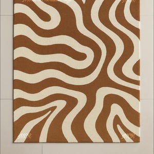 May include: Brown and cream-colored rug with a wavy pattern. The rug has a square shape and features a repeating design of flowing, organic lines. The text "Modern Creation Rugs" is visible on the top of the rug. The letters "MCR" are visible in the corners.