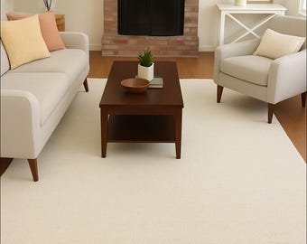 May include: A cream-colored rectangular rug in a living room setting. The rug is placed on a hardwood floor, with a sofa, armchair, and coffee table. The room features a fireplace and a large plant. The rug has a textured surface.