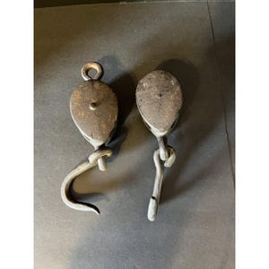 May include: Two antique metal pulleys with hooks. The pulleys are made of a dark, weathered metal, showing signs of age and use. Each pulley has a rounded body with a central pivot point and a curved hook attached below. The hooks are also made of metal.