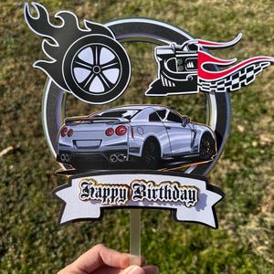 May include: A silver car cake topper with a banner that reads "Happy Birthday". The topper features a race car, a wheel with flames, and a checkered flag design. The background is green.