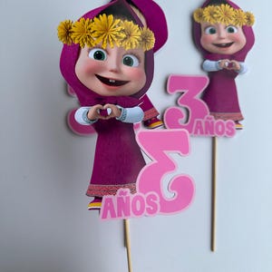 Pode incluir: Duas decora&ccedil;&otilde;es de bolo com uma personagem de desenho animado com um vestido e um chap&eacute;u roxos, com uma coroa de flores amarelas. O n&uacute;mero "3" e a palavra "A&Ntilde;OS" em rosa tamb&eacute;m est&atilde;o inclu&iacute;dos. As decora&ccedil;&otilde;es s&atilde;o presas a palitos de madeira.