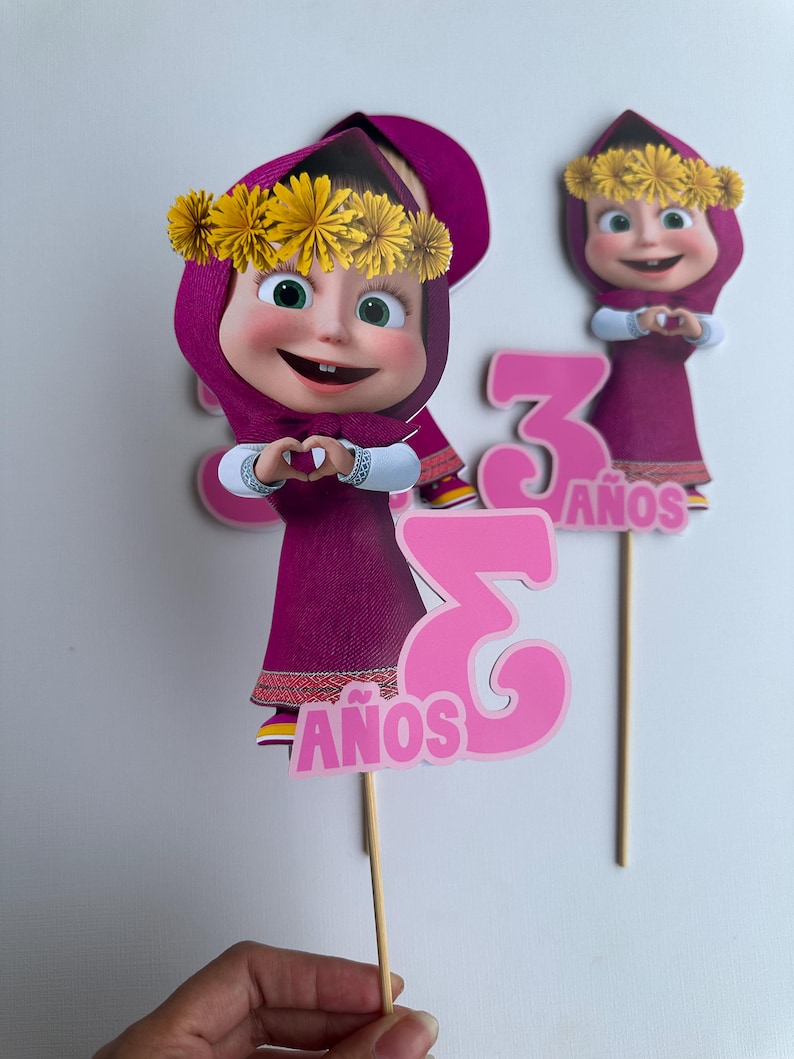 Pode incluir: Duas decora&ccedil;&otilde;es de bolo com uma personagem de desenho animado com um vestido e um chap&eacute;u roxos, com uma coroa de flores amarelas. O n&uacute;mero "3" e a palavra "A&Ntilde;OS" em rosa tamb&eacute;m est&atilde;o inclu&iacute;dos. As decora&ccedil;&otilde;es s&atilde;o presas a palitos de madeira.