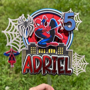 May include: A colorful cake topper featuring Spider-Man in a dynamic pose, with the number 5 and the name "ADRIEL". The design includes spiderwebs, a cityscape, and a red, blue, and black color scheme. The topper is designed for a birthday celebration.
