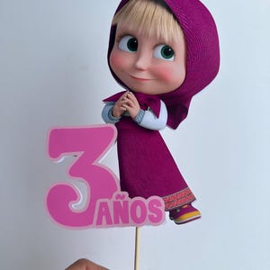 Pode incluir: Topper de bolo com uma personagem de desenho animado com vestido e capuz roxos, com o n&uacute;mero 3 e a palavra "ANOS" em rosa. A personagem tem olhos verdes e uma express&atilde;o sorridente. O topper &eacute; segurado por um palito de madeira.