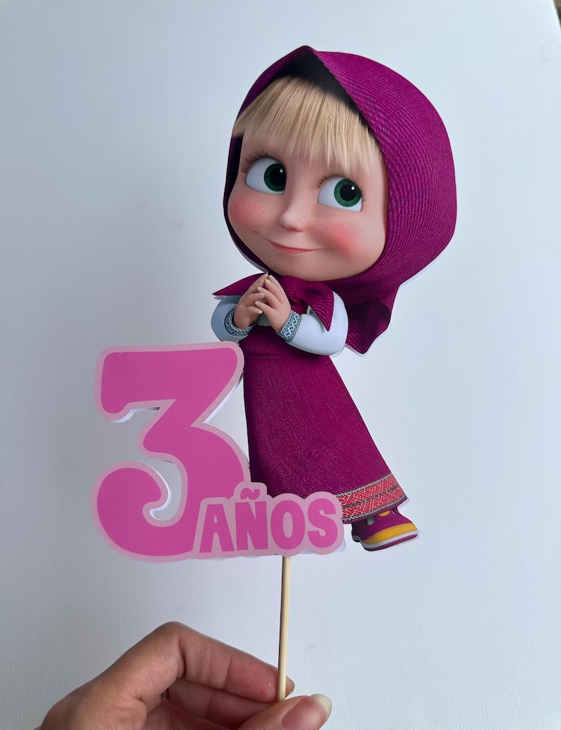 Pode incluir: Topper de bolo com uma personagem de desenho animado com vestido e capuz roxos, com o n&uacute;mero 3 e a palavra "ANOS" em rosa. A personagem tem olhos verdes e uma express&atilde;o sorridente. O topper &eacute; segurado por um palito de madeira.
