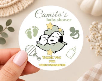 May include: A round white sticker featuring the text Camila's baby shower and THANK YOU FOR YOUR PRESENCE. The design includes a black and white dog sleeping on a sage green cloud, surrounded by yellow stars, sage green baby footprints, a pacifier, a rattle, and a baby bottle.
