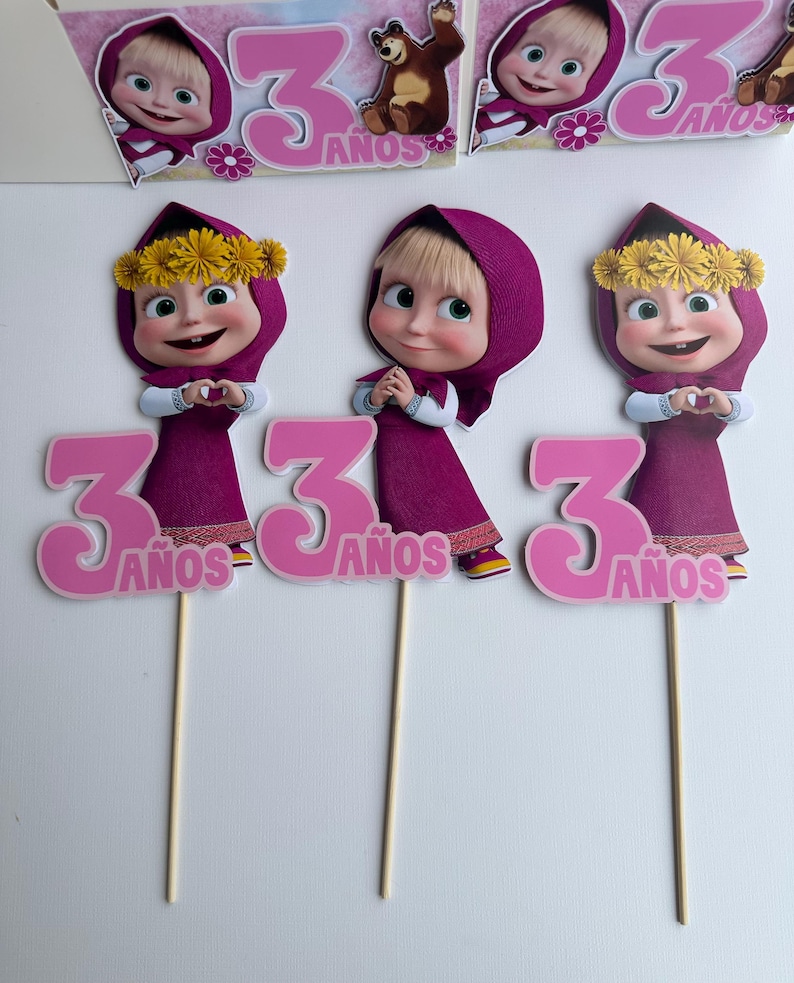 Pode incluir: Tr&ecirc;s topos de bolo com uma menina de desenho animado em um vestido e capuz roxos, com uma coroa de flores amarelas. Cada topo tem o n&uacute;mero 3 rosa e a palavra "A&ntilde;os". Os topos est&atilde;o em palitos de madeira.