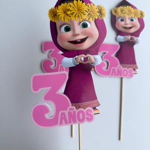 Pode incluir: Topper de bolo rosa e roxo com um personagem de desenho animado com uma coroa de flores, formando um cora&ccedil;&atilde;o com as m&atilde;os. O n&uacute;mero "3" e a palavra "A&ntilde;os" tamb&eacute;m s&atilde;o vis&iacute;veis.
