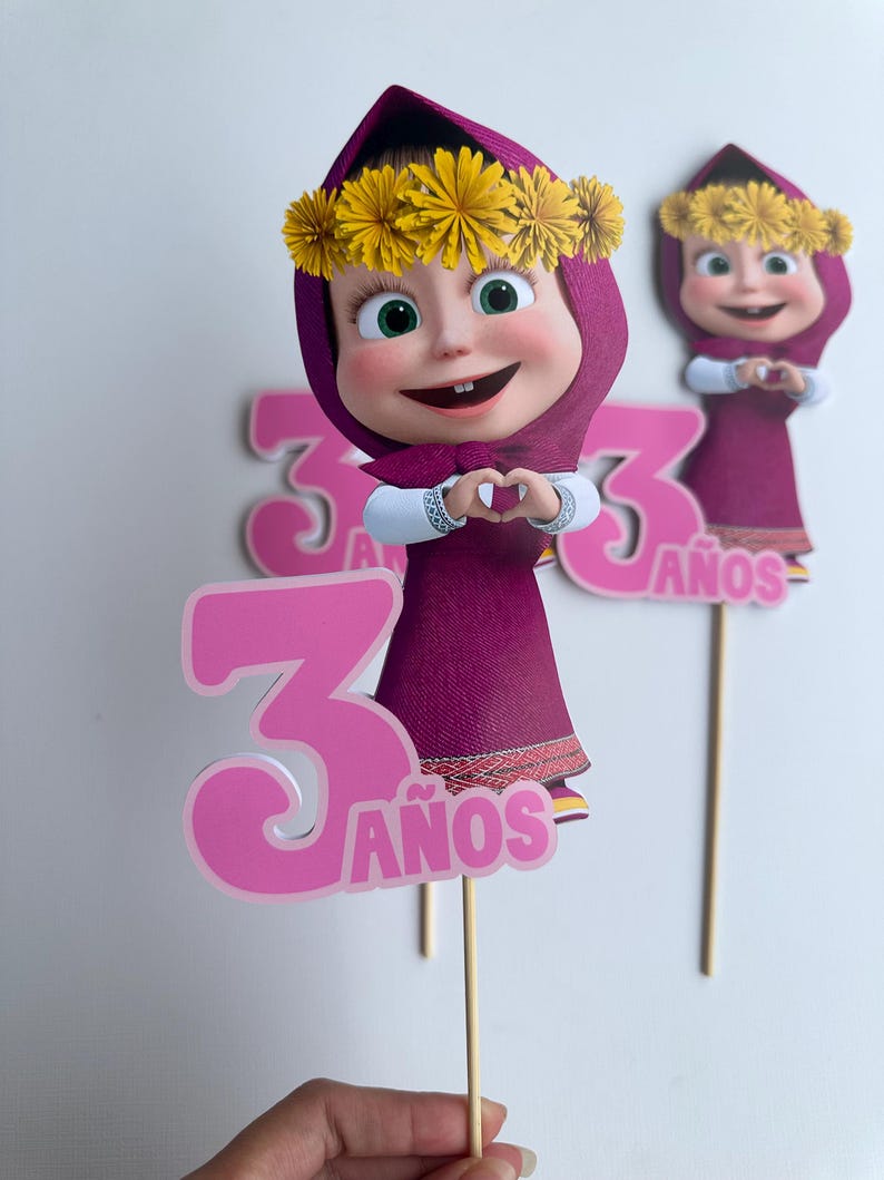 Pode incluir: Topper de bolo rosa e roxo com um personagem de desenho animado com uma coroa de flores, formando um cora&ccedil;&atilde;o com as m&atilde;os. O n&uacute;mero "3" e a palavra "A&ntilde;os" tamb&eacute;m s&atilde;o vis&iacute;veis.