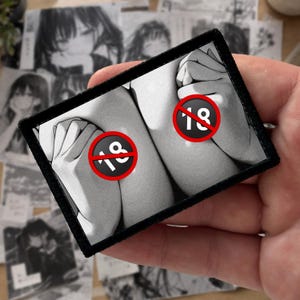 May include: A rectangular embroidered patch featuring a black and white manga-style illustration of two hands covering a chest area. The design includes two circular red and white warning symbols with the number 18 printed in the centre of each.