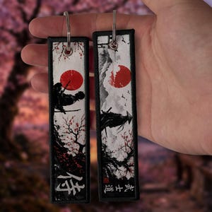 May include: Two rectangular fabric keychains with black stitched borders. Each features a white background with a red sun, a black silhouette of a samurai figure, and cherry blossom branches. The left tag displays the kanji 侍 and the right tag displays 武士道.