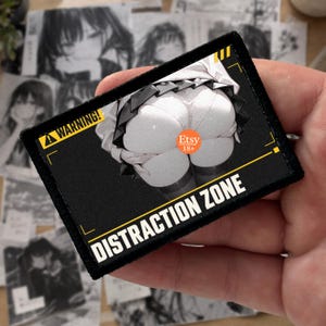May include: A rectangular black patch with a white graphic illustration of a person wearing a pleated skirt. The patch features yellow corner accents, a yellow warning sign with the text WARNING, and the white text DISTRACTION ZONE.