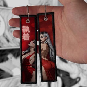 Pode incluir: Dois porta-chaves retangulares de tecido com um fundo vermelho vibrante e a ilustração de uma pessoa com cabelo comprido prateado e um vestido vermelho. O porta-chaves da esquerda exibe caracteres kanji brancos.
