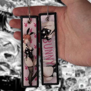 Pode incluir: Dois porta-chaves retangulares de tecido com bordas costuradas em preto e argolas de metal prateado. O fundo rosa claro apresenta um logótipo de coelho vermelho repetido. Um porta-chaves mostra uma personagem anime em body preto, o outro diz BUNNY.