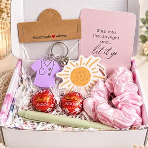 Könnte beinhalten: Eine Geschenkbox mit einem lila Schlüsselanhänger in Form eines Kasacks, einem sonnenförmigen Aufkleber mit dem Text YOU ARE LOVED, einer rosa Karte mit dem Text Step into the daylight and.. let it go, zwei roten Lindt Lindor Pralinen, einem rosa Haargummi und einem hellgrünen Stift.