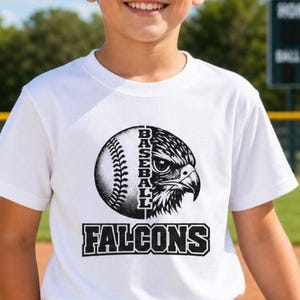 May include: A white short-sleeved t-shirt featuring a black graphic print on the chest. The design shows a baseball merged with a falcon head, the vertical text BASEBALL, and the word FALCONS printed in a bold, block font below the graphic.