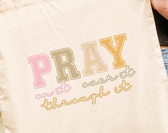 May include: A cream-coloured tote bag featuring the text "PRAY over it through it" in a retro font. The bag is held by a person wearing a brown jacket and blue jeans. The background is blurred with green foliage. The bag has long handles.