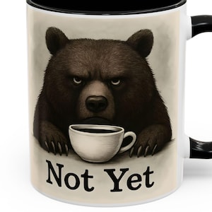 Può includere: Tazza da caffè in ceramica bianca con interno e manico neri. La tazza presenta un'illustrazione di un orso bruno arrabbiato che tiene una tazza di caffè, con le parole "Not Yet" stampate sotto.