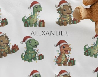 May include: White baby blanket featuring a repeating pattern of cartoon dinosaurs wearing Santa hats and holding gifts. The name "ALEXANDER" is printed in black. A tan teddy bear rests on the blanket.