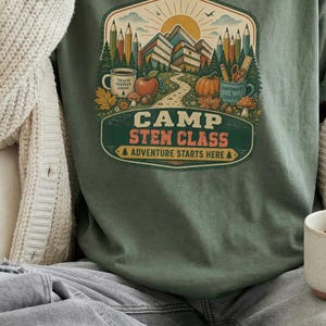 Peut inclure: Un t-shirt à manches courtes vert mousse avec une impression graphique représentant un paysage de montagne, des crayons, une tasse et une citrouille. Le texte indique CAMP STEM CLASS ADVENTURE STARTS HERE. Porté avec un gilet en maille crème.