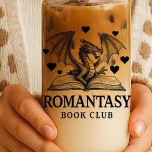 May include: A glass tumbler with a bamboo lid and straw, filled with a layered drink. The glass is decorated with a dragon illustration, the text "ROMANTASY BOOK CLUB", and black heart accents. The tumbler is held by someone wearing a cream-colored sweater.