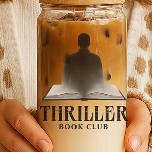 Op de afbeelding: Een glazen beker met een bamboe deksel en een rietje, gevuld met ijskoffie. De beker heeft een afbeelding van een persoon die op een open boek zit, met de woorden "THRILLER BOOK CLUB" in zwart en wit.