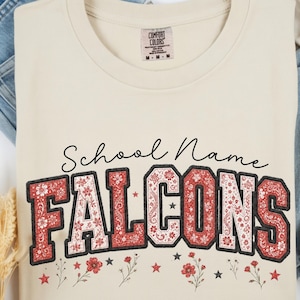 以下が含まれることがあります： クリーム色のTシャツで、黒い筆記体で「School Name」と書かれ、その下に大きなブロック体で「FALCONS」とプリントされています。文字は赤と白のフローラルペイズリー柄で黒い縁取りがあり、小さな赤い花と星の模様がアクセントになっています。