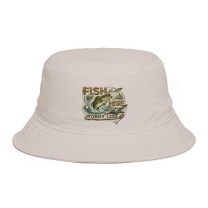 May include: A light beige bucket hat with a graphic featuring a jumping fish, the words "FISH MORE" and "WORRY LESS". The graphic is in shades of green, brown, and blue. The hat is a casual accessory.