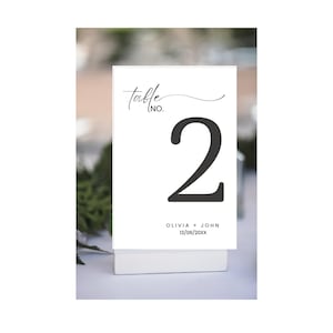 Può includere: Un cartoncino bianco con il numero del tavolo 2 in un carattere grigio scuro. La parola "table" è scritta sopra il numero in un carattere corsivo. I nomi "Olivia + John" e una data sono stampati in basso.