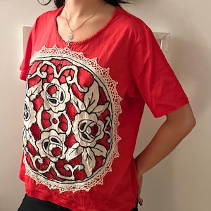 May include: A red short-sleeved t-shirt with a round floral design on the front. The design features red roses with white petals and black outlines, surrounded by a white lace trim. The shirt has a scoop neckline.