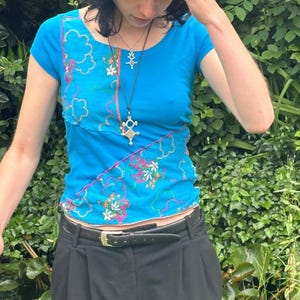 Peut inclure: Un haut bleu à manches courtes avec des motifs floraux et nuageux roses et blancs. Le haut a une encolure dégagée et est associé à un pantalon noir et une ceinture noire. Un collier pendentif argenté est visible.