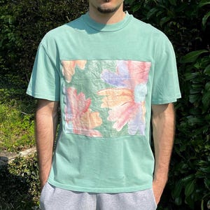 Può includere: Una maglietta verde menta con un motivo floreale. La maglietta presenta una toppa rettangolare sul davanti con un motivo floreale colorato nei toni del rosa, arancione e viola. Ha maniche corte e girocollo.