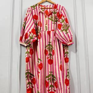 Peut inclure: Une longue robe à rayures verticales roses, blanches et beiges. La robe présente un imprimé floral de fleurs rouges avec des feuilles vertes. Elle a des manches longues et bouffantes et un col à nouer. La robe est suspendue à un cintre en bois.