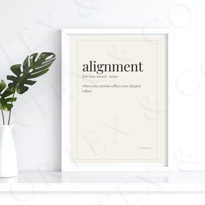 May include: A framed print with the word "alignment" and its definition. A white vase with green leaves is to the left of the print. The print is in a white frame.
