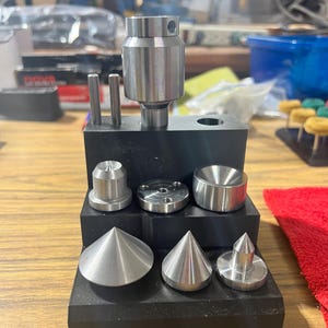 May include: A collection of metal tools and components, including cylindrical and conical shapes, arranged on a black base. The tools are made of shiny, silver-colored metal and appear to be precision-engineered.