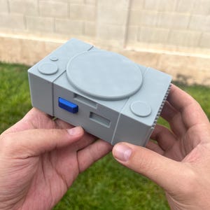 May include: A gray, 3D-printed model of a classic video game console, held in two hands. The model features a disc tray, buttons, and a power button. The console is a light gray color with a blue button. The model is a replica of a retro gaming console.