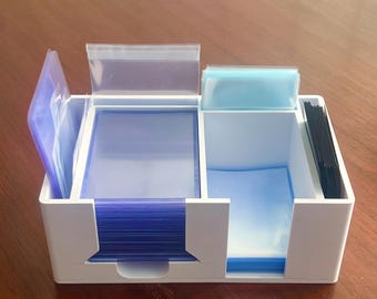 May include: A white plastic card holder with multiple compartments. The compartments contain stacks of blue and clear card sleeves, and black card dividers. The holder is designed to organize and store trading cards or similar items.